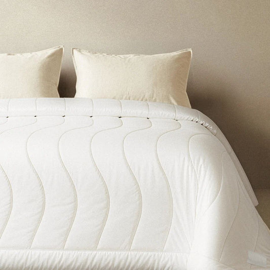 White Breeze Comforter laid over a bed, with two cream pillows against a warm beige wall.